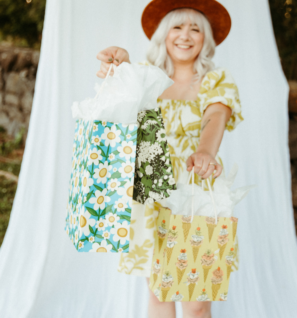 A woman is featured holding 3 gift bags closer to you. The gift bags are covered in retro florals, and colorful patterns. Greens, Blues, Yellows, and Orange colortones are all featured in these gift wrap bags. 